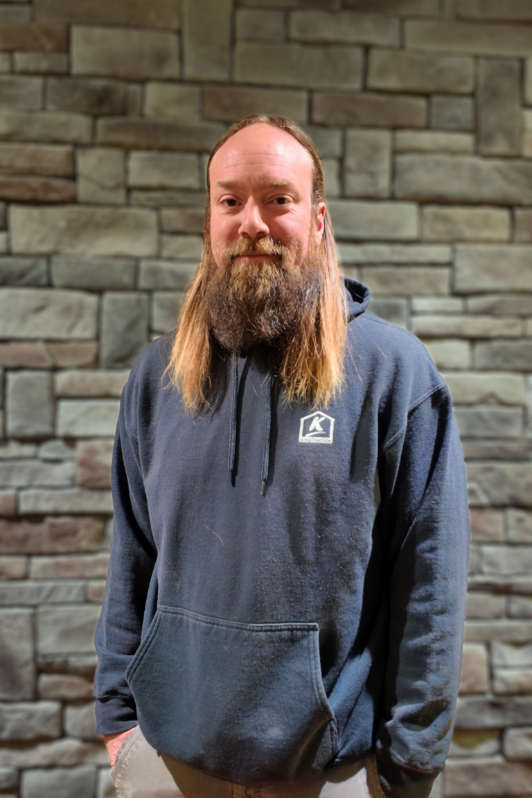 Man with long beard and mustache wearing a navy hoodie stands against a stone wall backdrop.