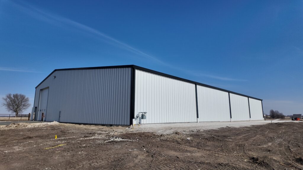 A newly constructed steel frame building with a dark gray roof and light gray siding, set against a backdrop of the rolling green Kansas Flint Hills under a clear blue sky.